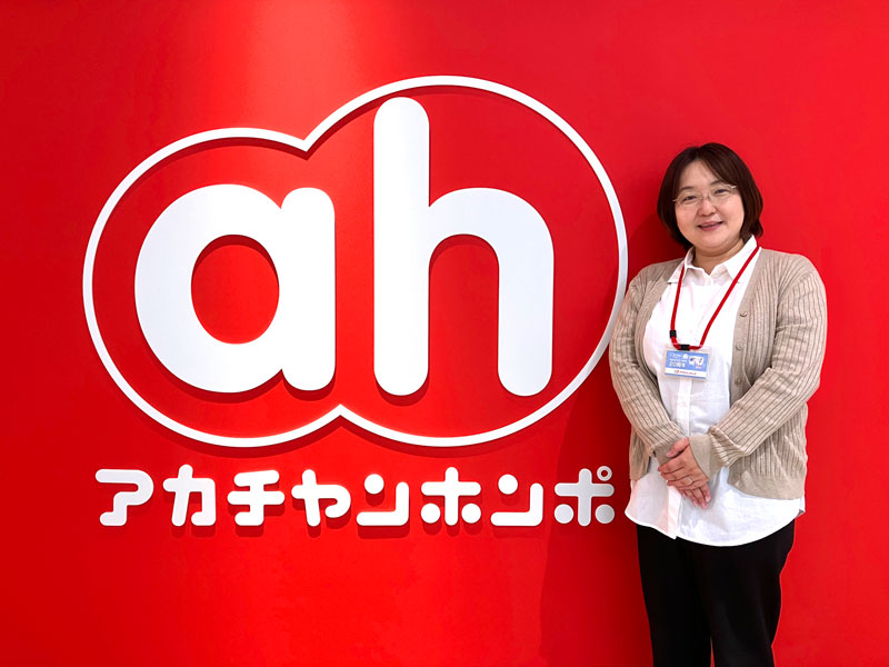Person standing beside a large red wall with a white 'ah' logo and Japanese text, wearing a name badge and cardigan.