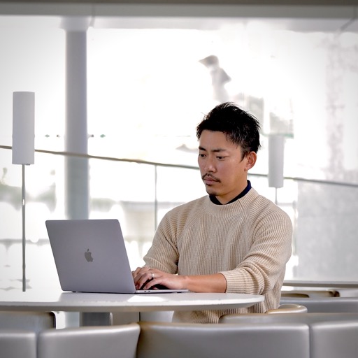 Man in a beige knit sweater sits at a round white table, typing on a silver laptop in a bright, modern space.