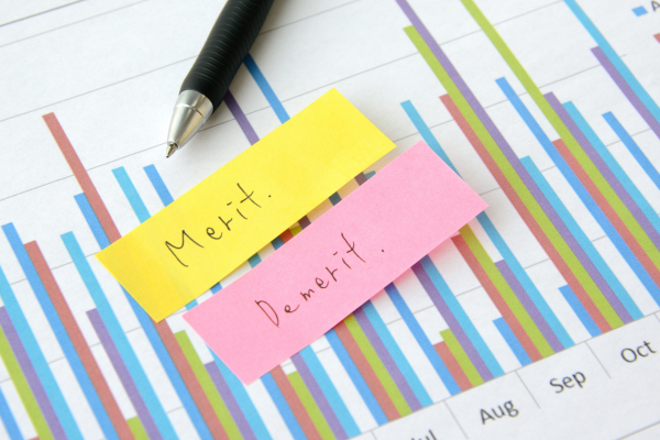 Close-up of a pen and two sticky notes placed on a colorful bar chart; yellow note says 'Meritx' and pink note says 'Dembrit' (handwritten names).メリット、デメリットと書かれた付箋メモ、棒グラフ