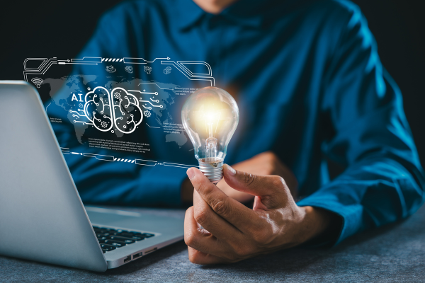 Person in a blue shirt holds a glowing light bulb beside a laptop, with a holographic AI brain graphic projected above the desk.
AIによりアイデアが浮かぶ、パソコン、電球