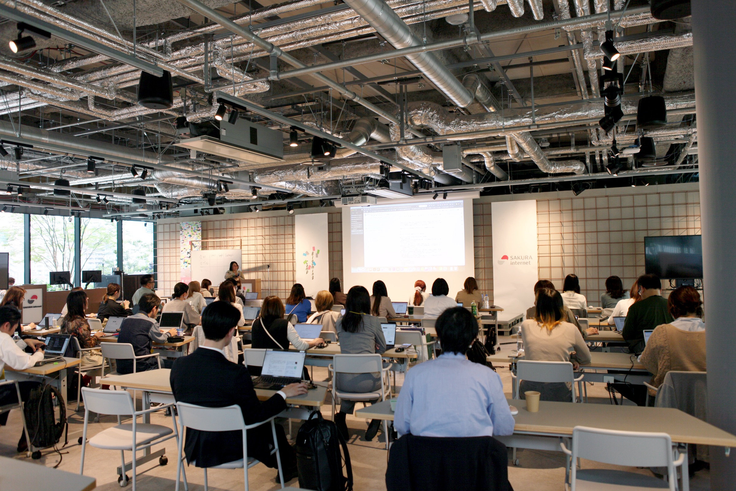 Conference room filled with people at desks using laptops, audience facing a projected presentation at the front, ceiling with exposed ducts visible in the background.