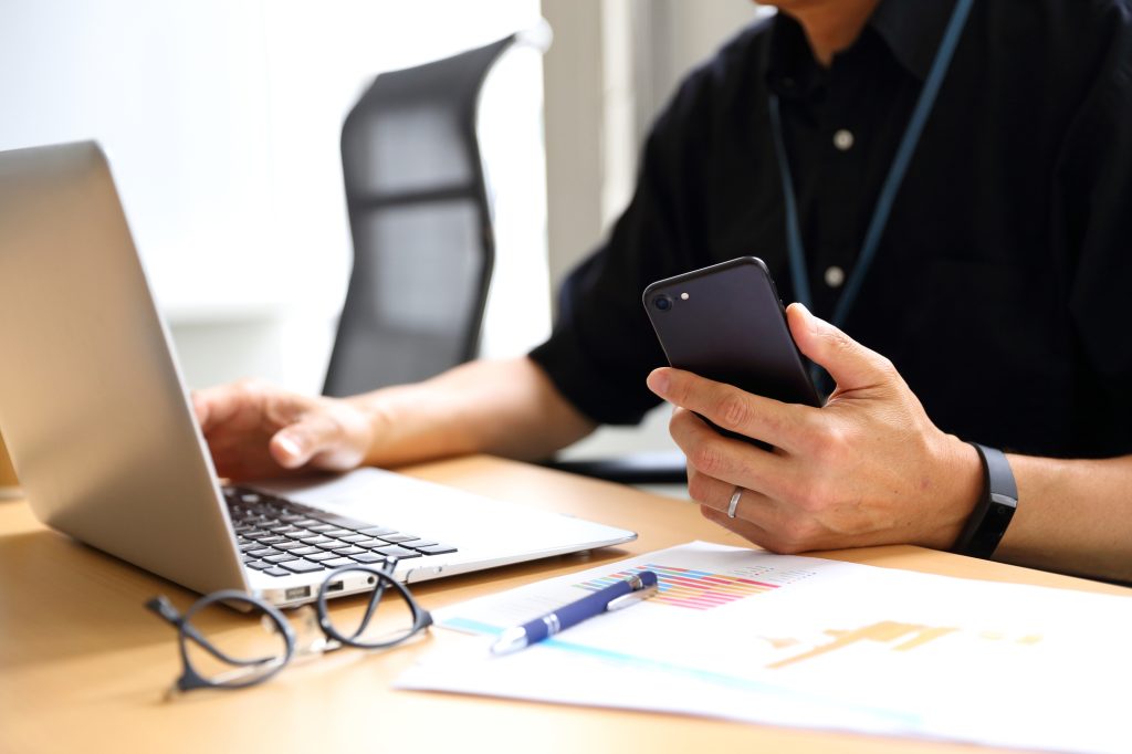 Person in a black shirt holds a smartphone while a laptop is open at a desk, with papers and glasses nearby.