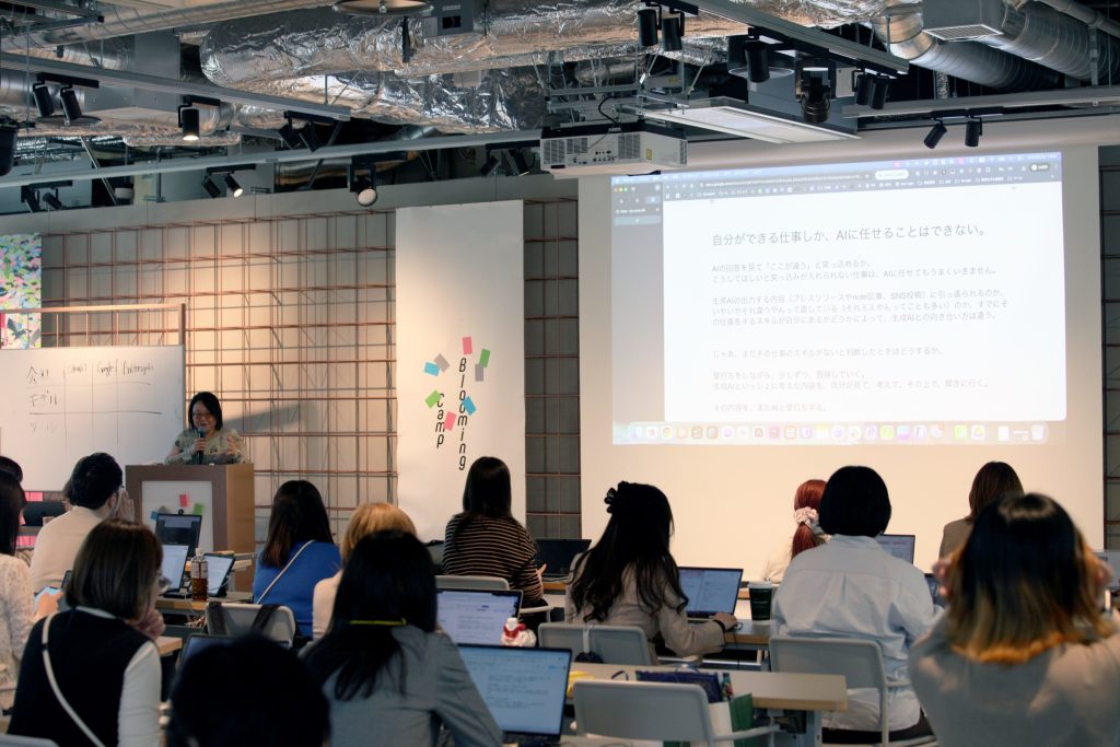 Speaker stands at a podium giving a presentation to a seated audience with laptops, projected slide visible on a large screen showing Japanese text.