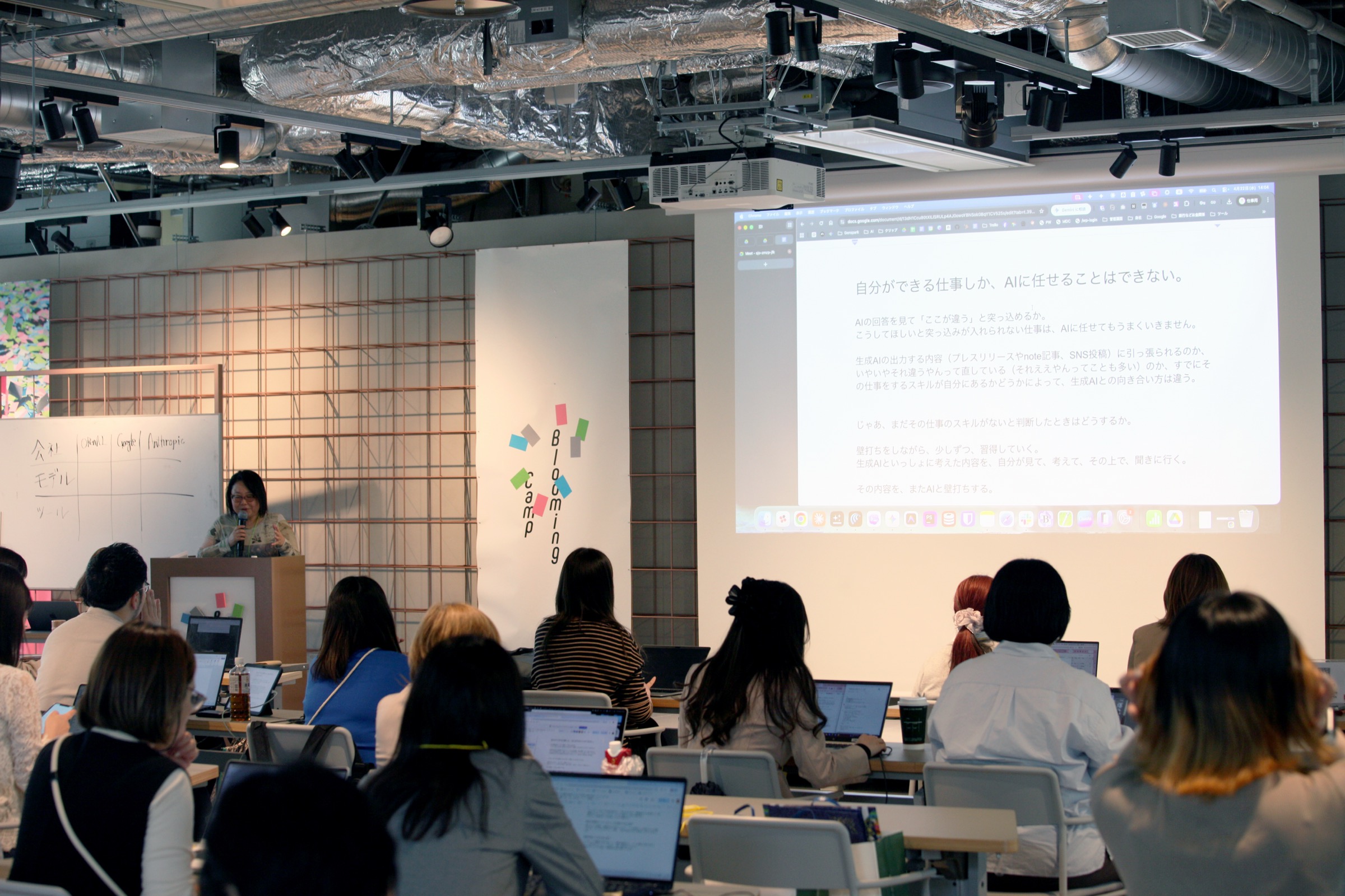 Speaker stands at a podium giving a presentation to a seated audience with laptops, projected slide visible on a large screen showing Japanese text.