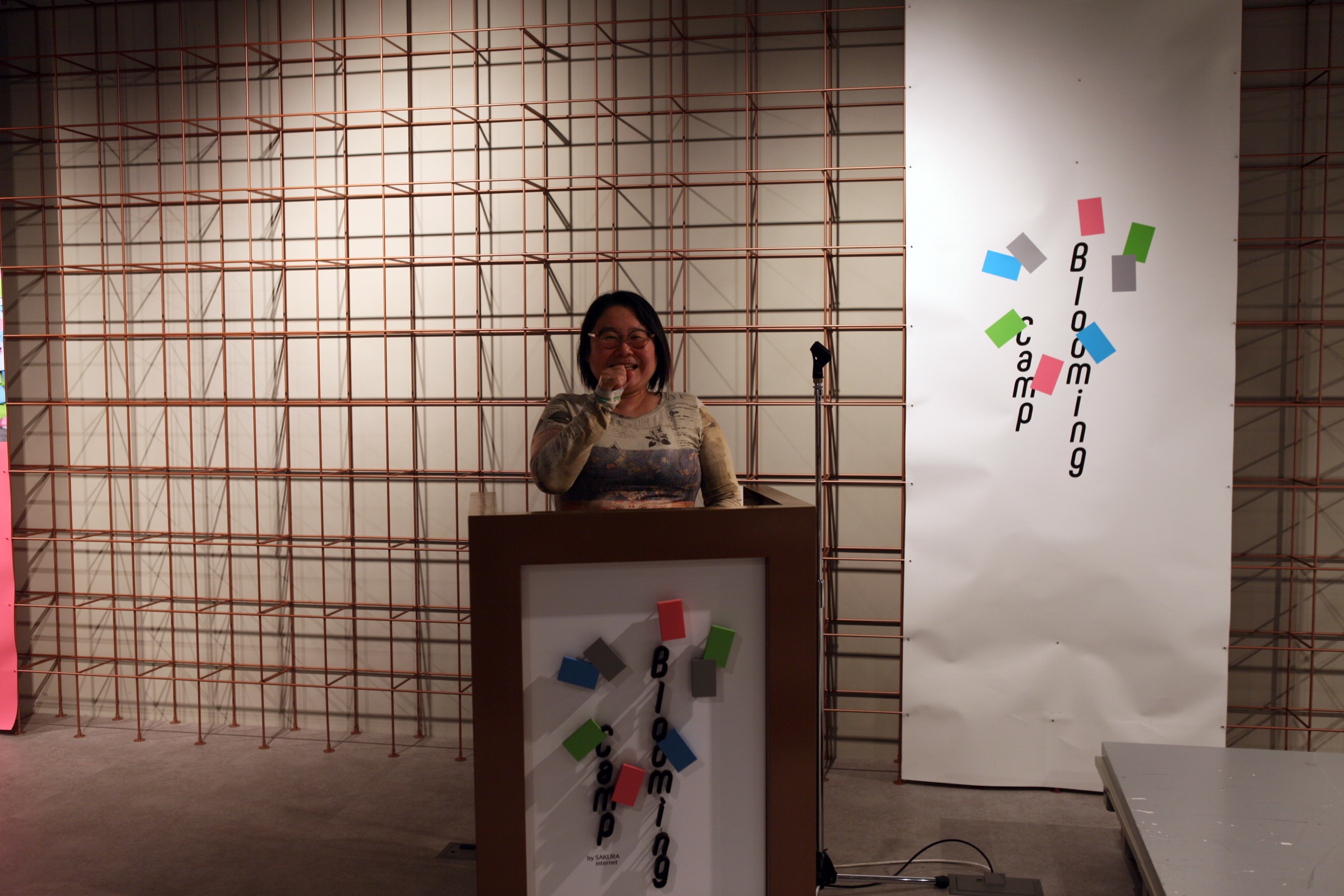 Smiling woman stands behind a wooden podium on a stage, with a copper grid backdrop and a white banner featuring colorful square shapes on the right side.