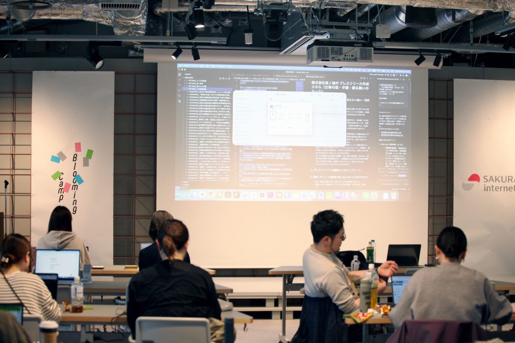 Conference room with participants seated at desks watching a large projection screen displaying a Japanese-language interface and code/texts on screen and banners on the sides.