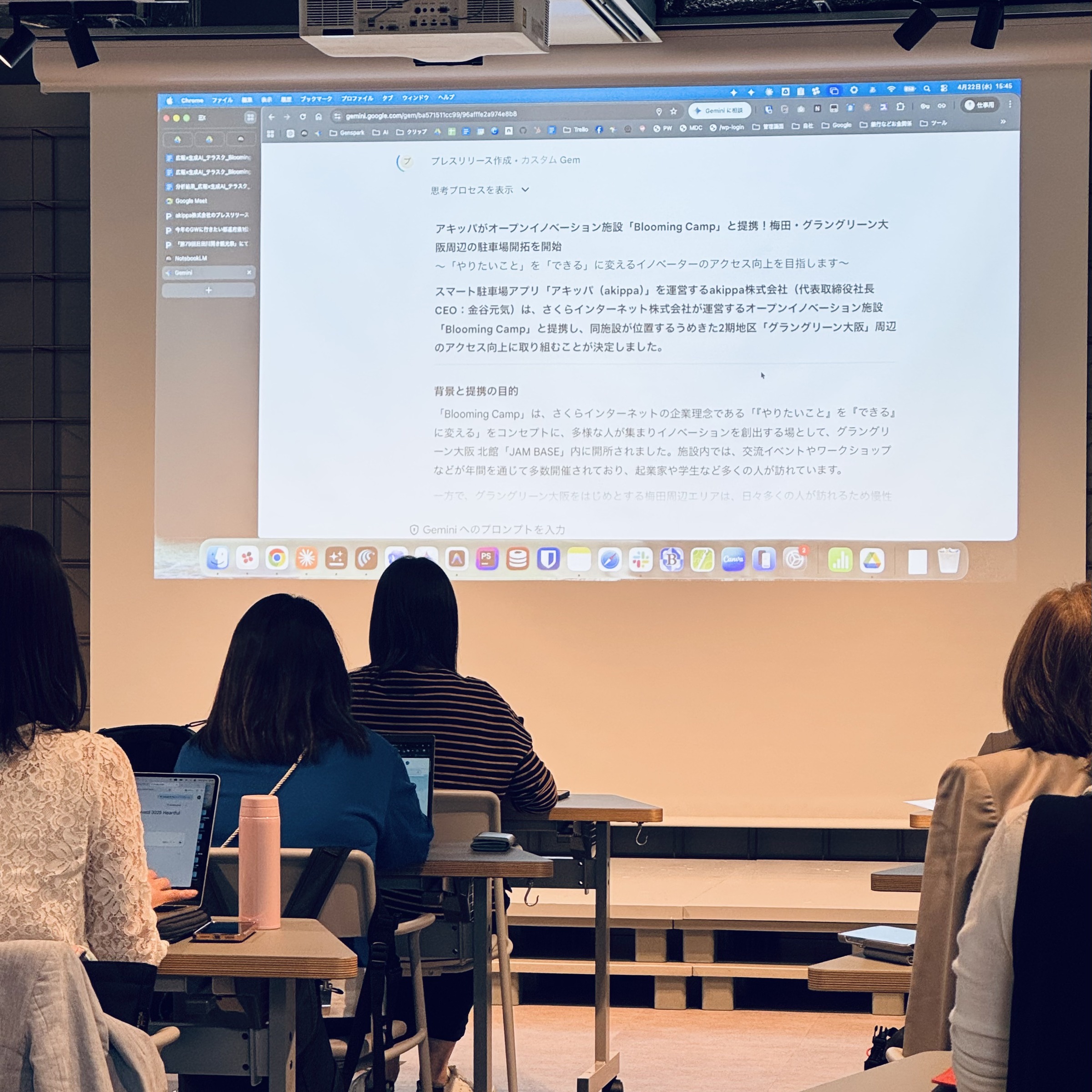 Audience seated at desks facing a large projection of a computer screen displaying Japanese text and a browser window, in a classroom setting.
