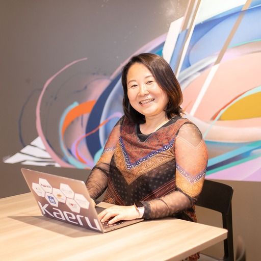 Smiling woman sits at a wooden table using a laptop with stickers, with a colorful abstract mural in the background.