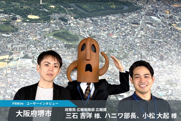Two men in suits pose with a tall brown cartoon mascot against a cityscape backdrop; a banner underneath reads Osaka Prefecture and Sakai City with interview host names.