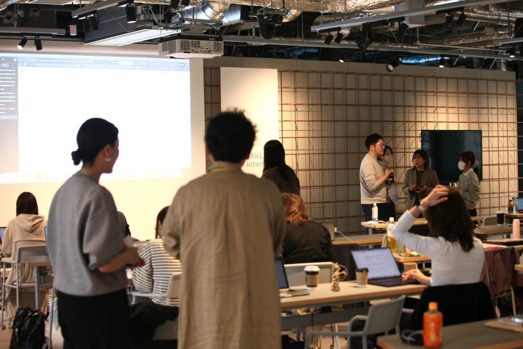 Group of people standing and talking at the front of a classroom-style room, with laptops on tables and a large projection screen visible on the left.