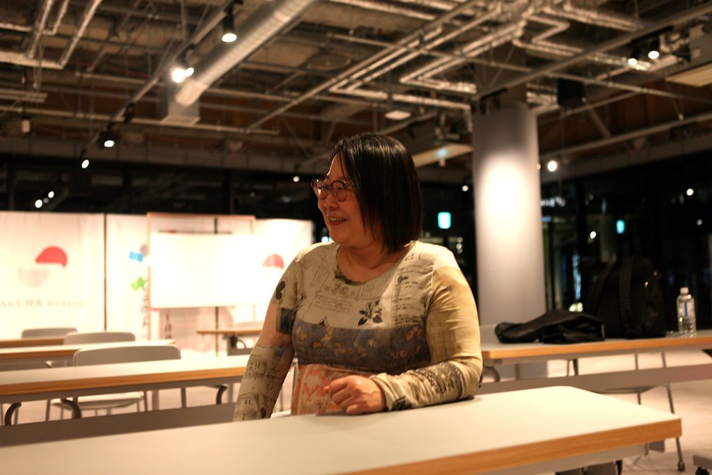 Woman with glasses smiling, leaning on a white table in a bright indoor event space.