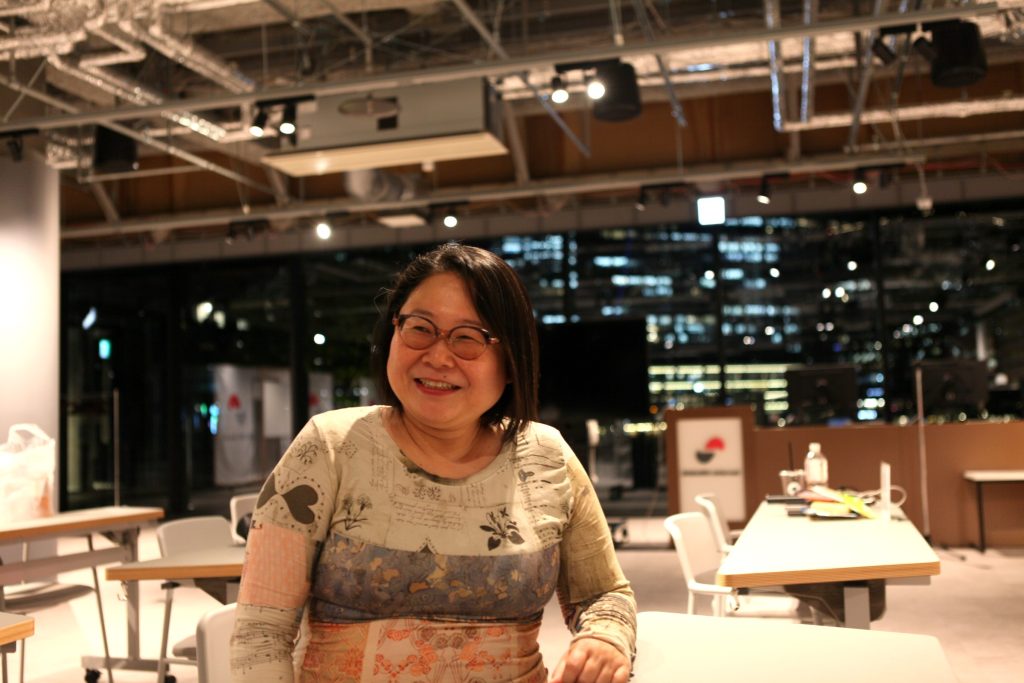 Smiling woman seated at a white table in a bright, modern workspace with office lights and a city view outside the windows.
