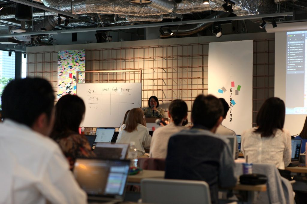 Speaker presenting at the front of a classroom-style conference room with a whiteboard and attendees using laptops in the foreground.