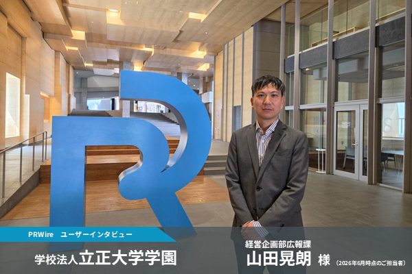A man in a gray suit stands beside a large blue abstract logo in a modern university lobby; interview feature banner at the bottom edge.