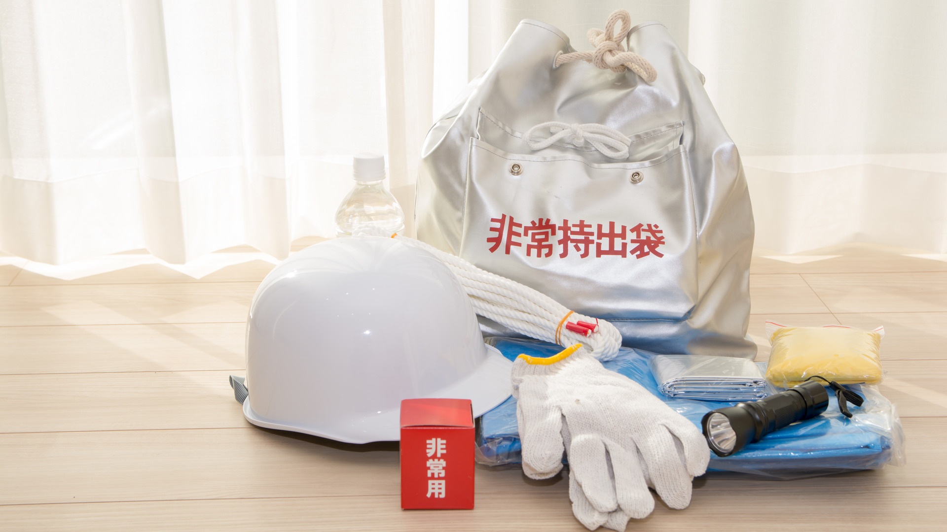 床に置かれた非常持出袋と、ヘルメット、水のボトル、ロープ、軍手、懐中電灯などの備え