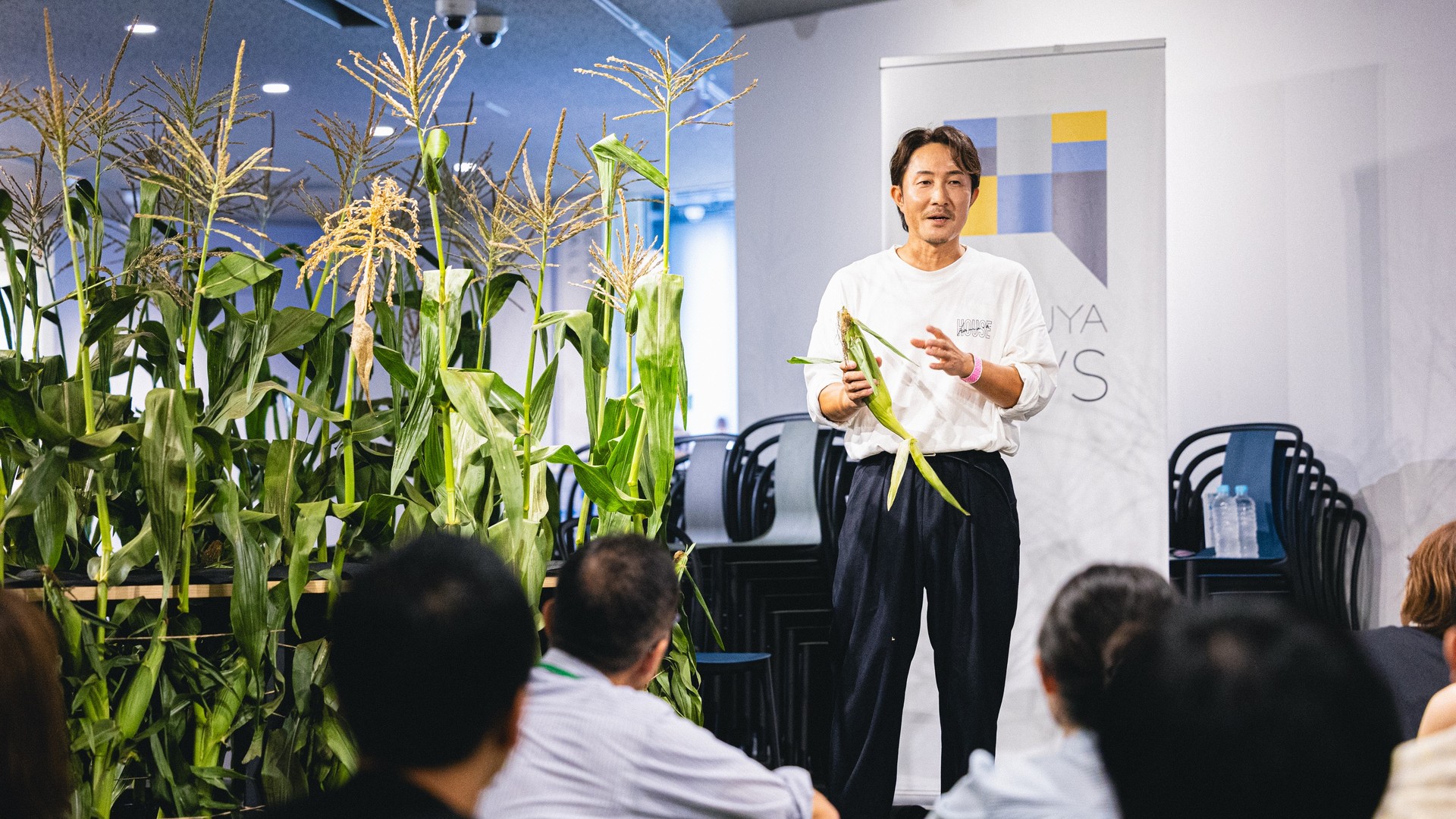 室内につくられたとうもろこし畑のわきで、とうもろこしを手にした男性が話している。手前に参加者の後ろ姿。
