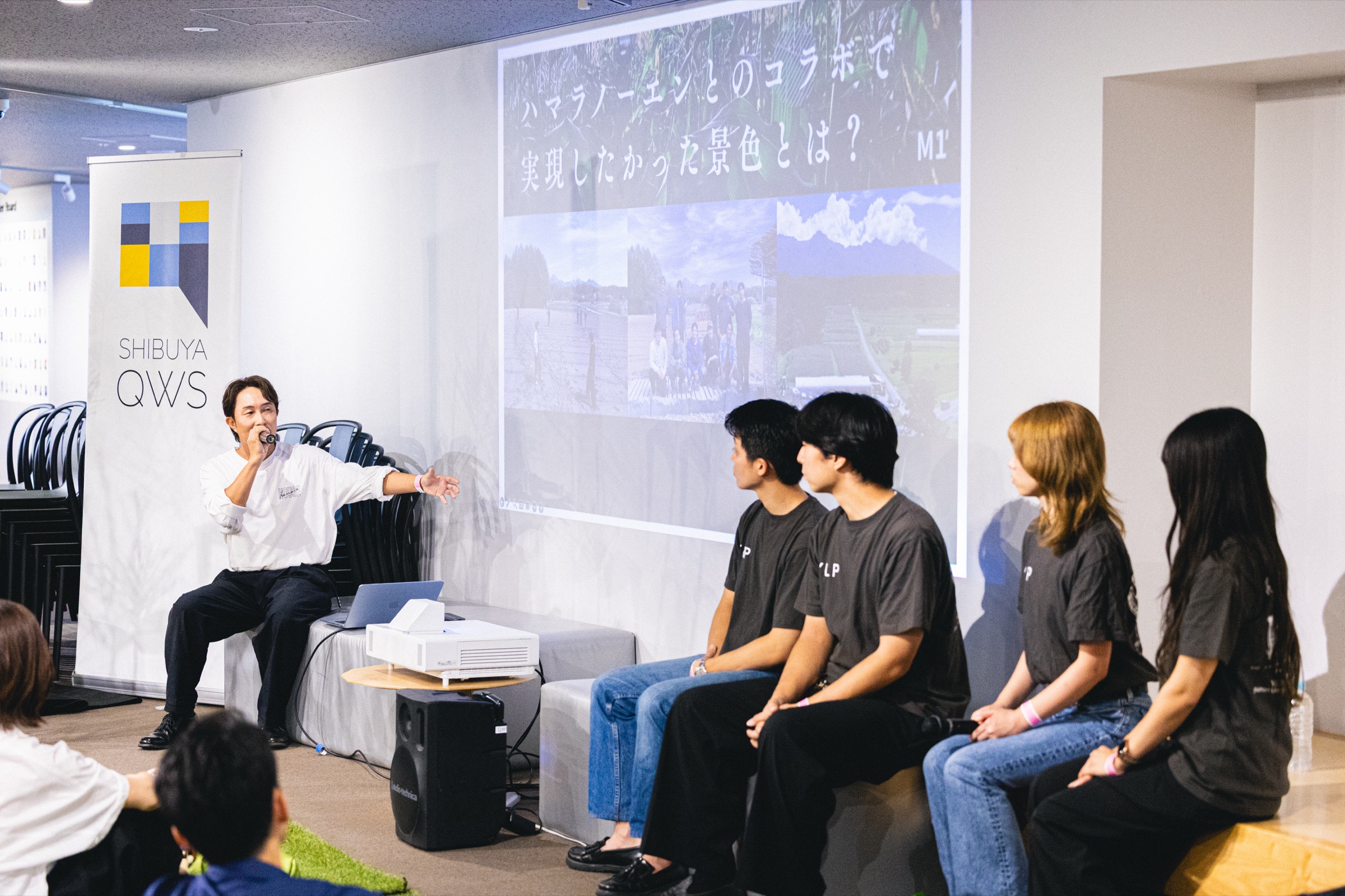 スクリーンの前に並んで座る4人の学生と、マイクを持って説明する男性。スクリーンには畑の写真が映っている。