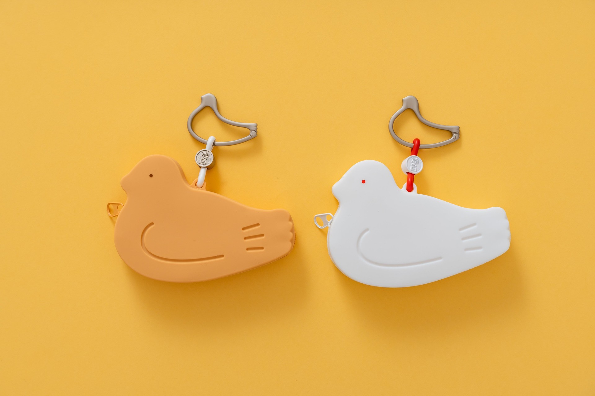 「鳩の日」限定シリコンポーチ「はとっこ」。鳩サブレーを思わせる茶と、ロゴをモチーフにした白。それぞれに鳩サブレーをあしらったカラビナが付く
