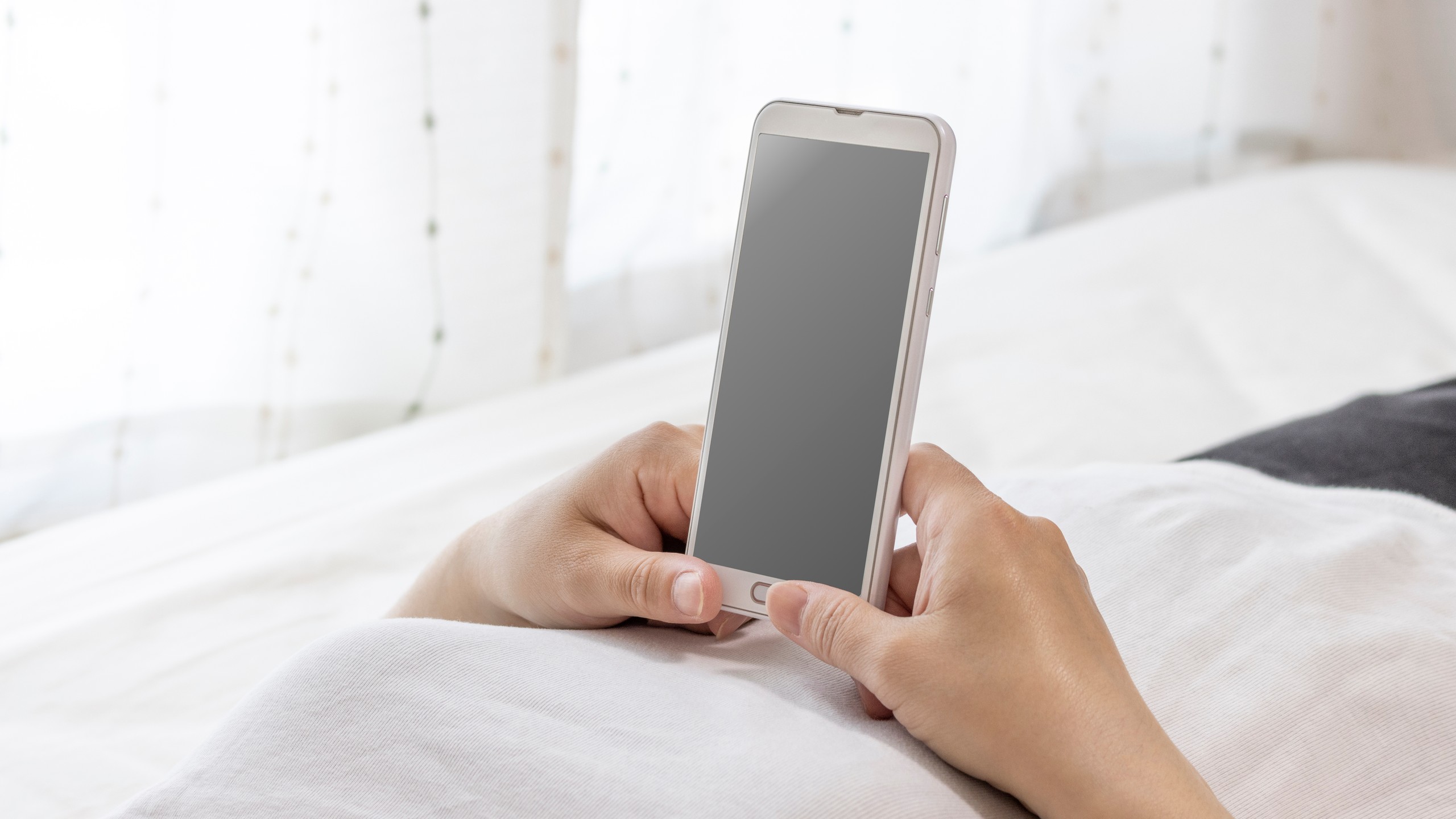 布団に横になり、両手でスマートフォンを操作する人の手元。画面には何も表示されていない