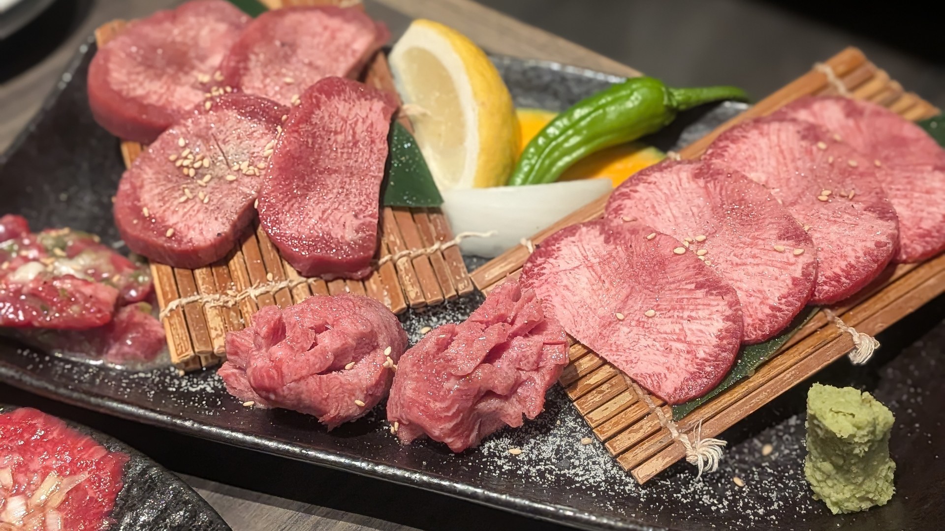 黒い長皿に、厚切りの牛タンと薄切りの肉が竹の敷き台に並べて盛られている。中央にレモンとしし唐、右下にわさびが添えられている。