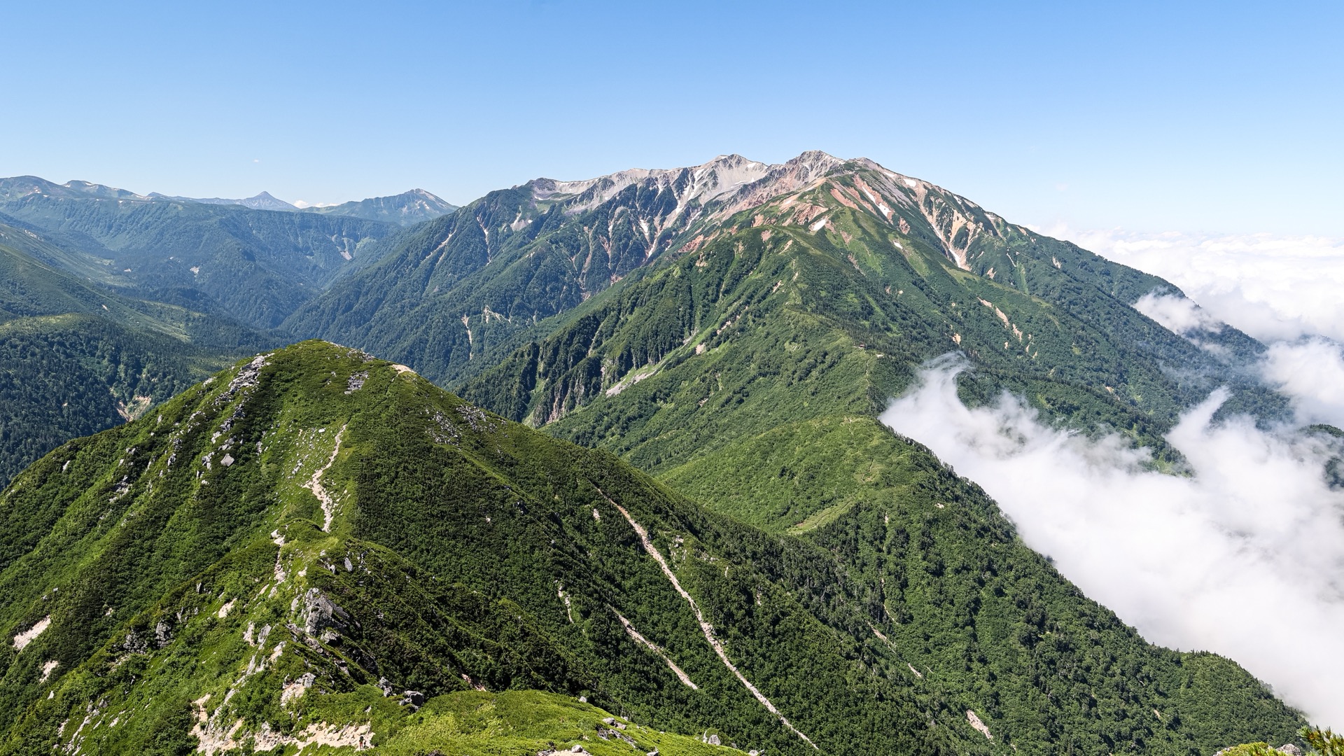 緑に覆われた夏山の稜線が手前から奥へ続き、雪渓の残る山並みの右手の谷に雲がかかっている風景